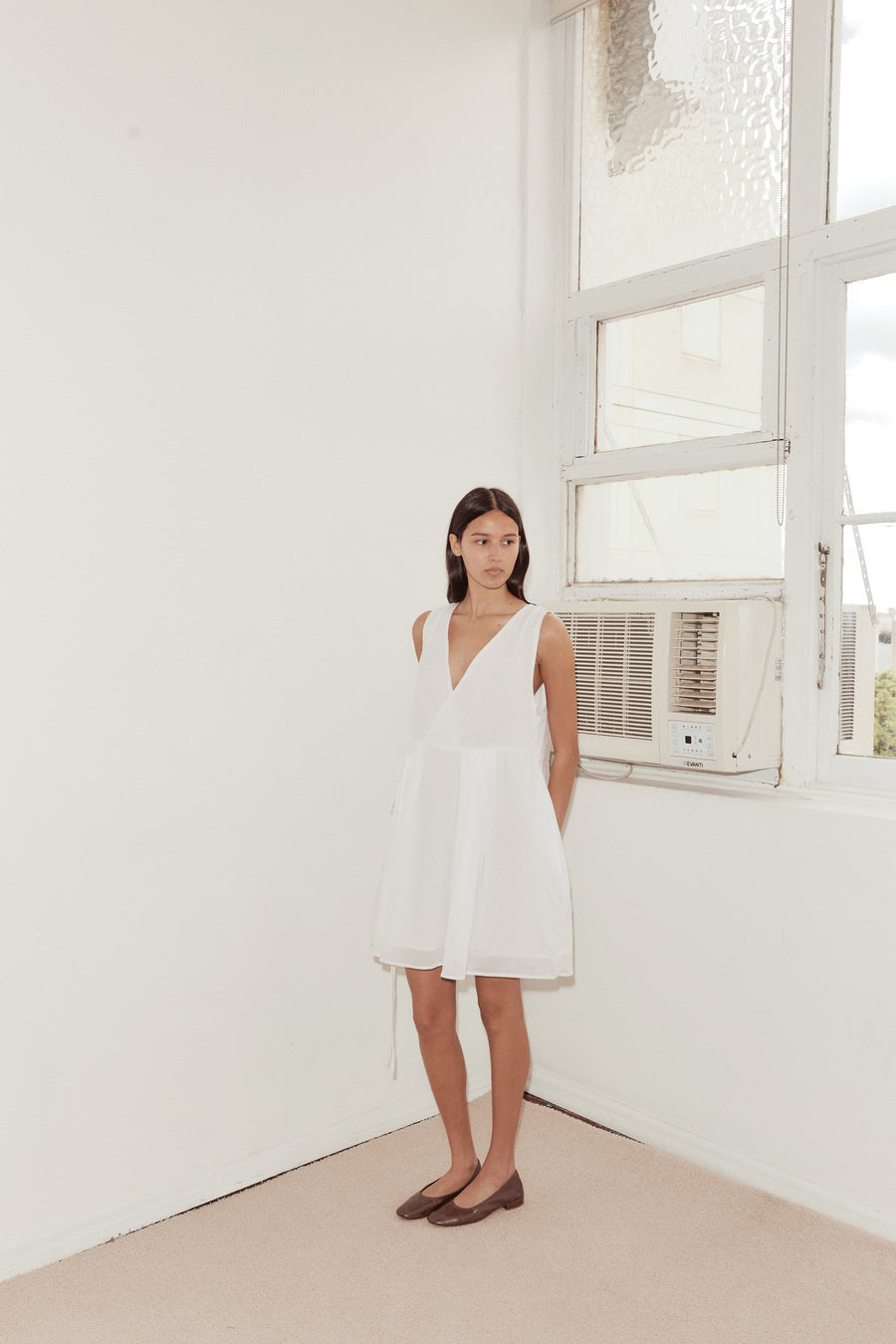 Long shot of female model standing against a white wall with a vintage airconditioning unit and dappled window wearing the Wrap Tie Dress in white by Deiji Studios. A relaxed airy mini dress with cross front v neckline, waist tie and double lined skirt.