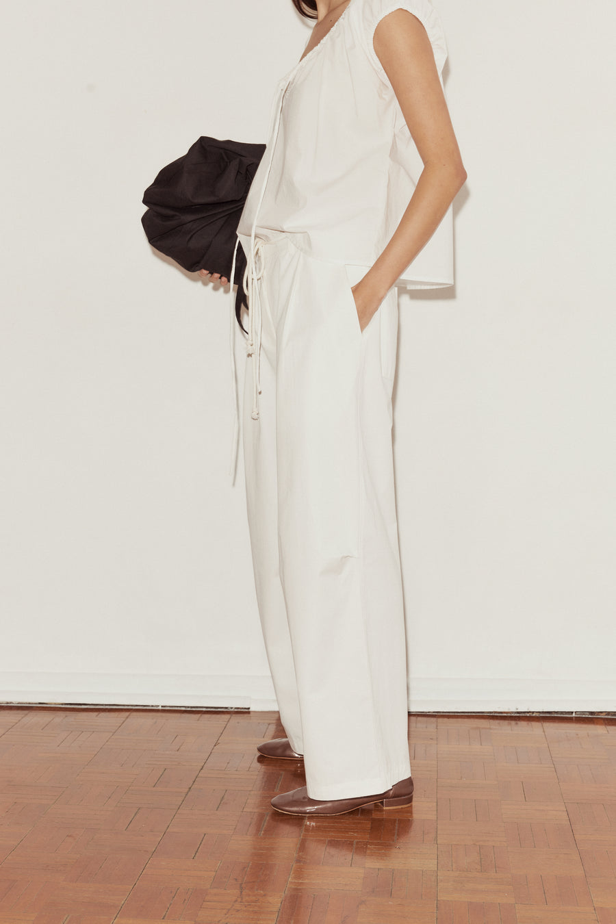 side shot of a female model wearing white Deiji Studios cotton twill pants, features a drawcord waist, deep side pockets and a relaxed leg in a full length. Styled with the Deiji Studios One Panel Top in white, features capped sleeves and a soft gathered neckline