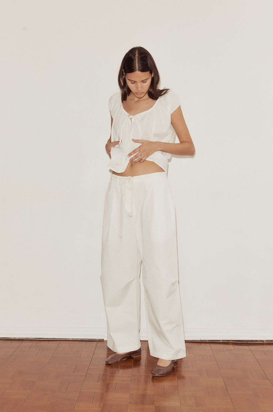 mid shot of a female model wearing white Deiji Studios cotton twill pants, features a drawcord waist and relaxed leg in a full length. Styled with the Deiji Studios One Panel Top in white, features capped sleeves and a soft gathered neckline