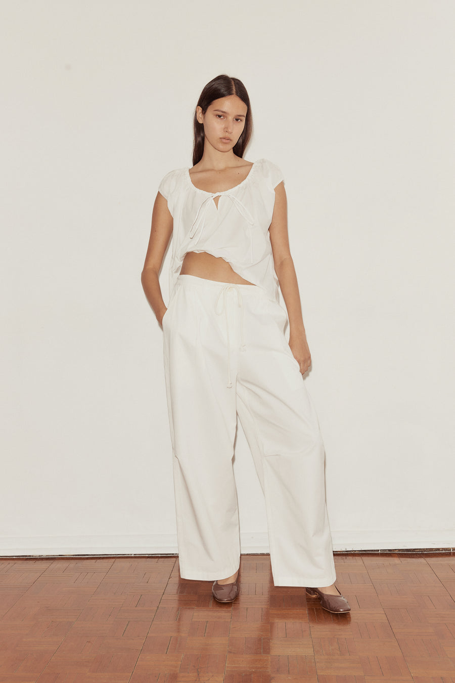mid shot of a female model wearing white Deiji Studios cotton twill pants, features a drawcord waist and relaxed leg in a full length. Styled with the Deiji Studios One Panel Top in white, features capped sleeves and a soft gathered neckline.