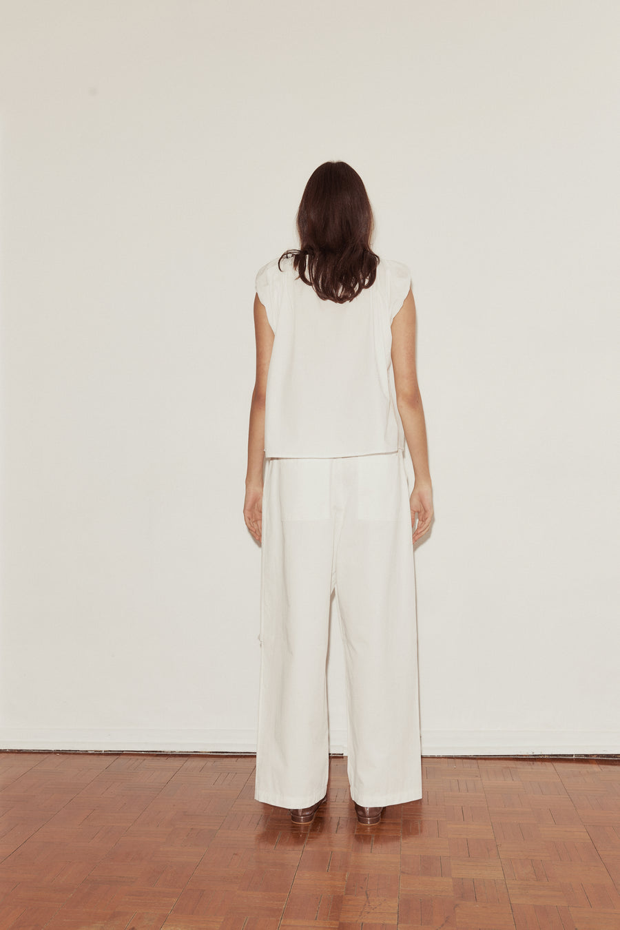 back shot of a female model wearing white Deiji Studios cotton twill pants, features a drawcord waist, deep back pockets and a relaxed leg in a full length. Styled with the Deiji Studios One Panel Top in white, features capped sleeves and a mid length