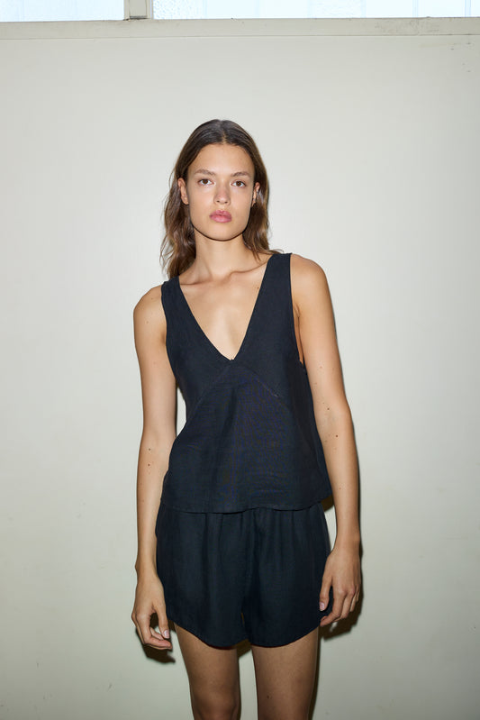 A woman with light brown hair and a neutral expression stands against an off-white wall, wearing Deiji Studios’ The Two Tie Set – Black: a sleeveless V-neck top and matching black shorts, her arms relaxed by her sides.