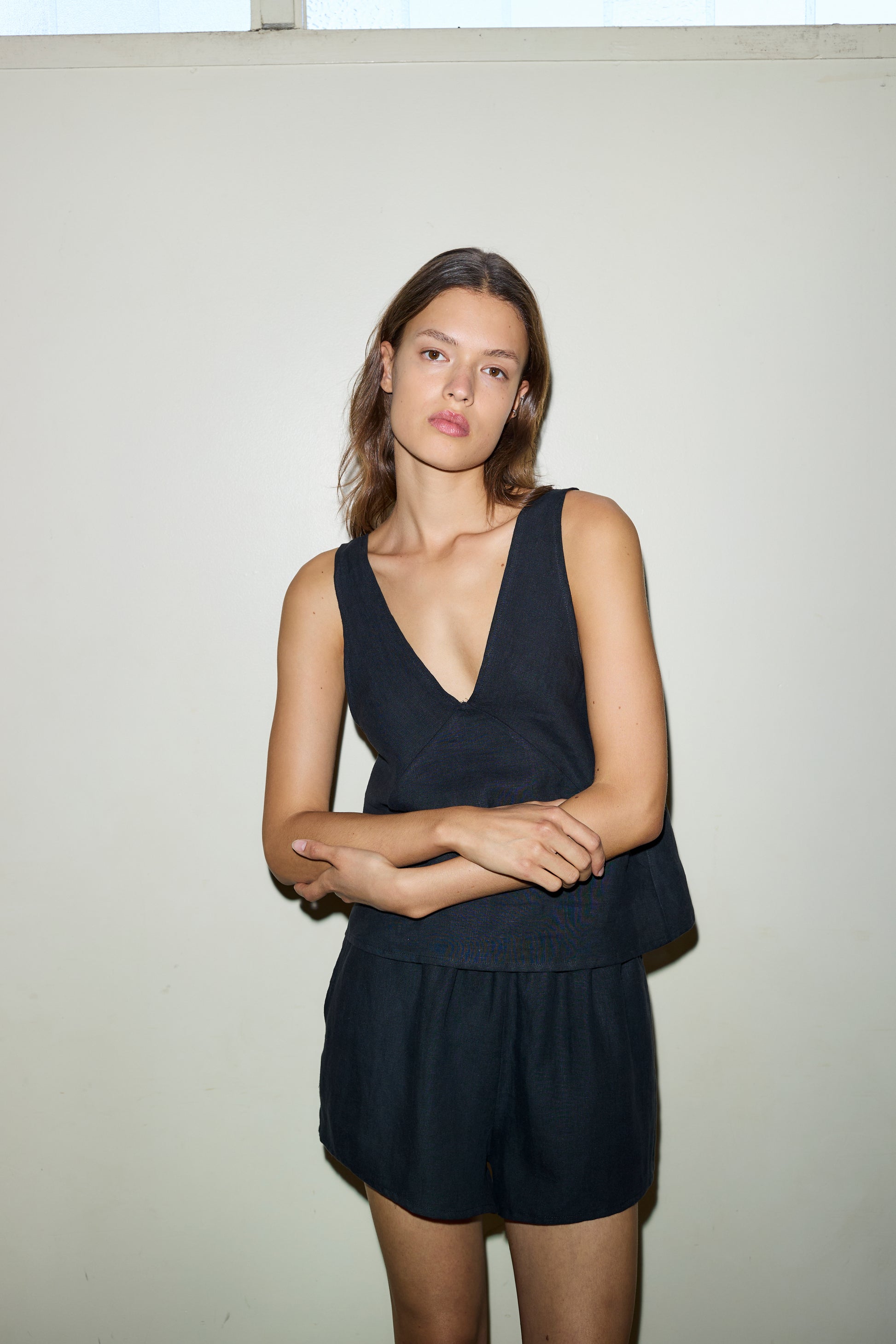 A woman with light brown hair and a neutral expression poses against a plain light wall, wearing The Two Tie Set - Black, which features a sleeveless deep V-neck French linen top and matching shorts. Her arms are loosely crossed.