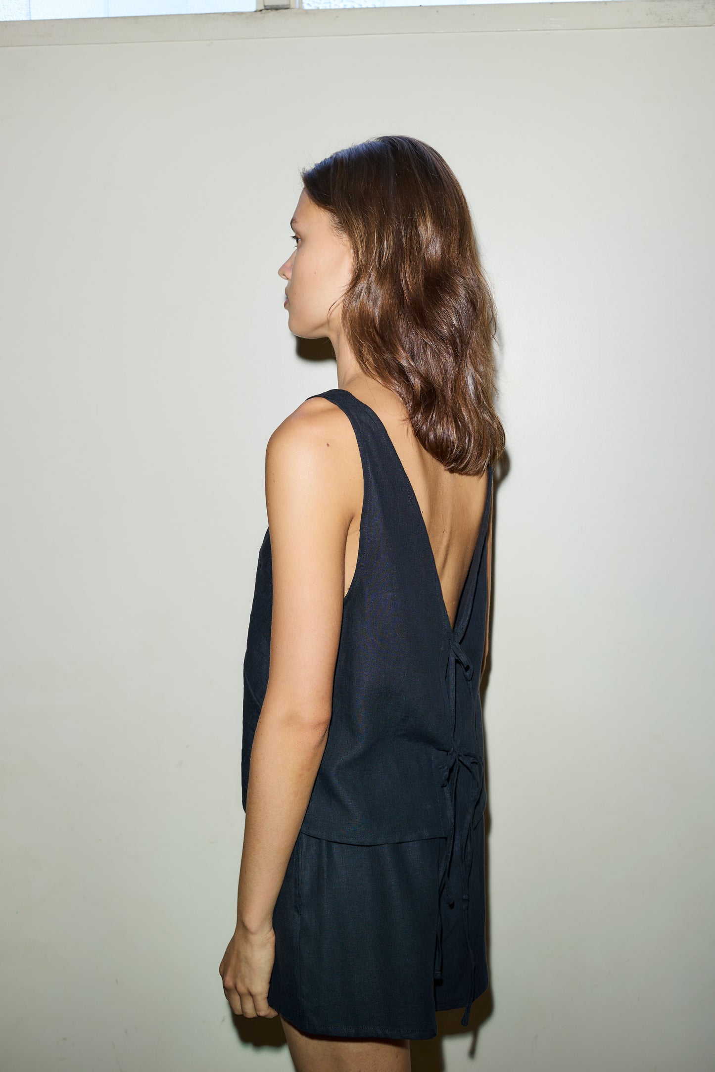 A woman with shoulder-length brown hair stands sideways against a plain light wall, wearing a sleeveless black "The Two Tie Set" by Deiji Studios featuring an open back with tie details.