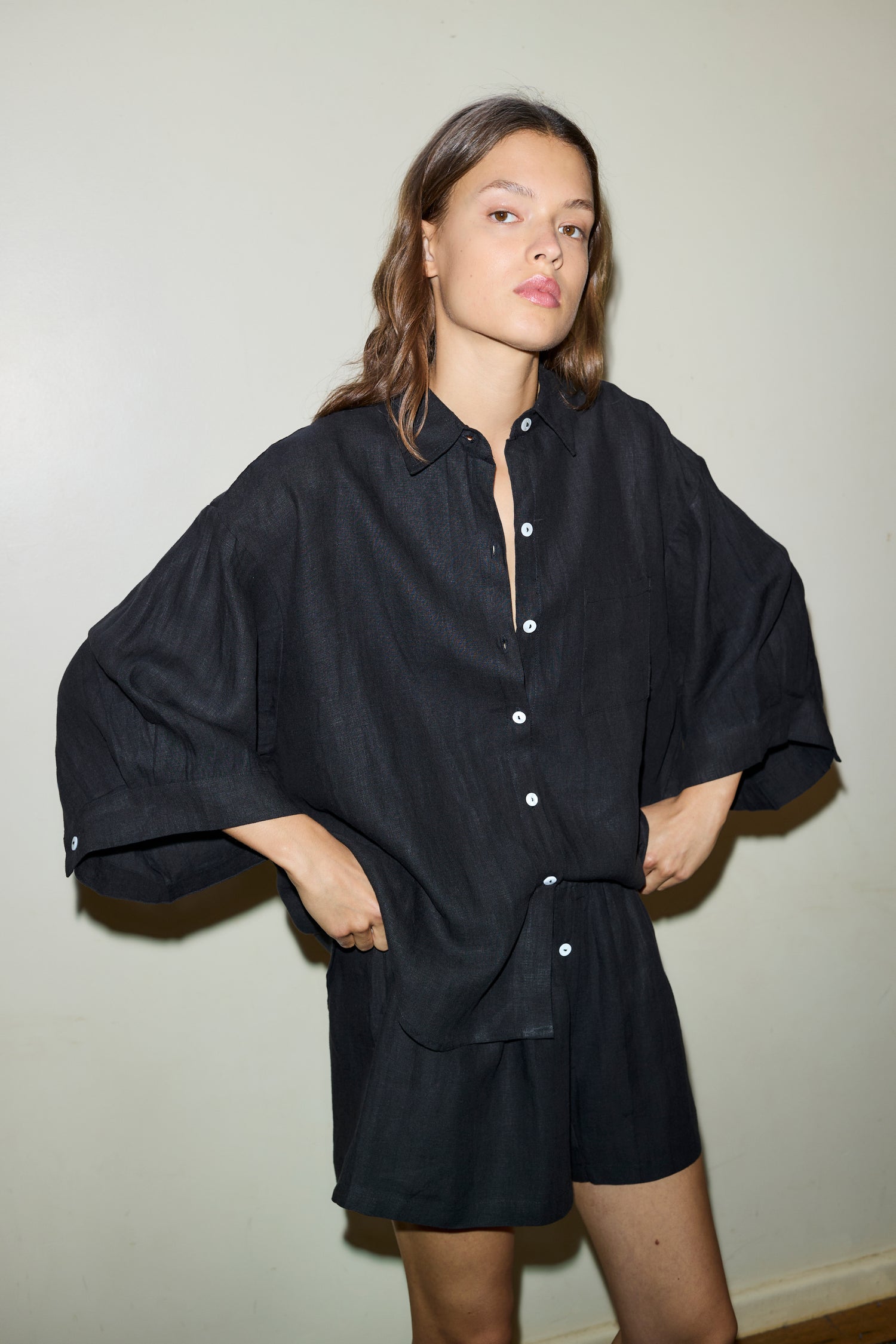 A person stands against a plain light-colored wall, wearing Deiji Studios’ the 03 set in deep black—an oversized linen shirt with rolled-up sleeves and matching boxer-style shorts—hands on hips, looking confidently at the camera.