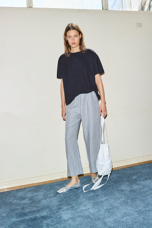 A woman with shoulder-length hair and a neutral expression stands indoors on blue carpet, wearing a loose black t-shirt, silver flats, a white drawstring bag, and the linen ease trouser - field check.