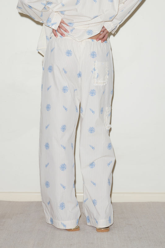 A person wearing the Cotton Pyjama Set - Sea Flower, featuring loose white organic cotton pants with blue floral and patchwork details, stands against a light background with hands on hips.