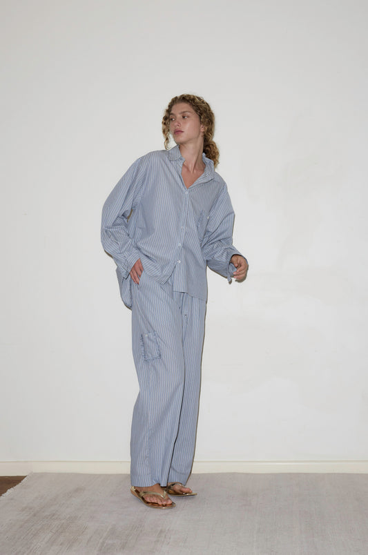 A person with curly hair stands indoors against a plain white wall, wearing the Cotton Pyjama Set - Glacier Stripe by Deiji Studios, with hands in pockets and gazing slightly to the side.