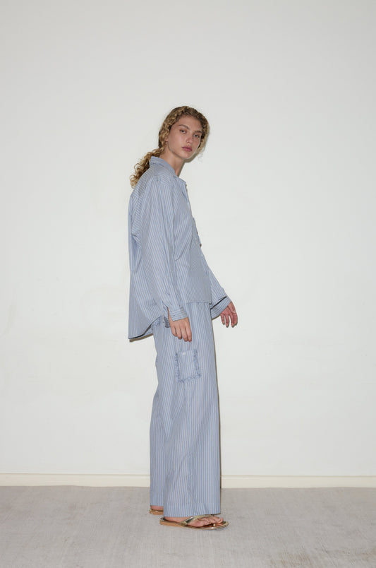 A person with curly hair stands sideways against a plain white wall, wearing the Cotton Pyjama Set - Glacier Stripe, a loose-fitting light blue and white striped ensemble with matching wide-leg pants and flip-flops.