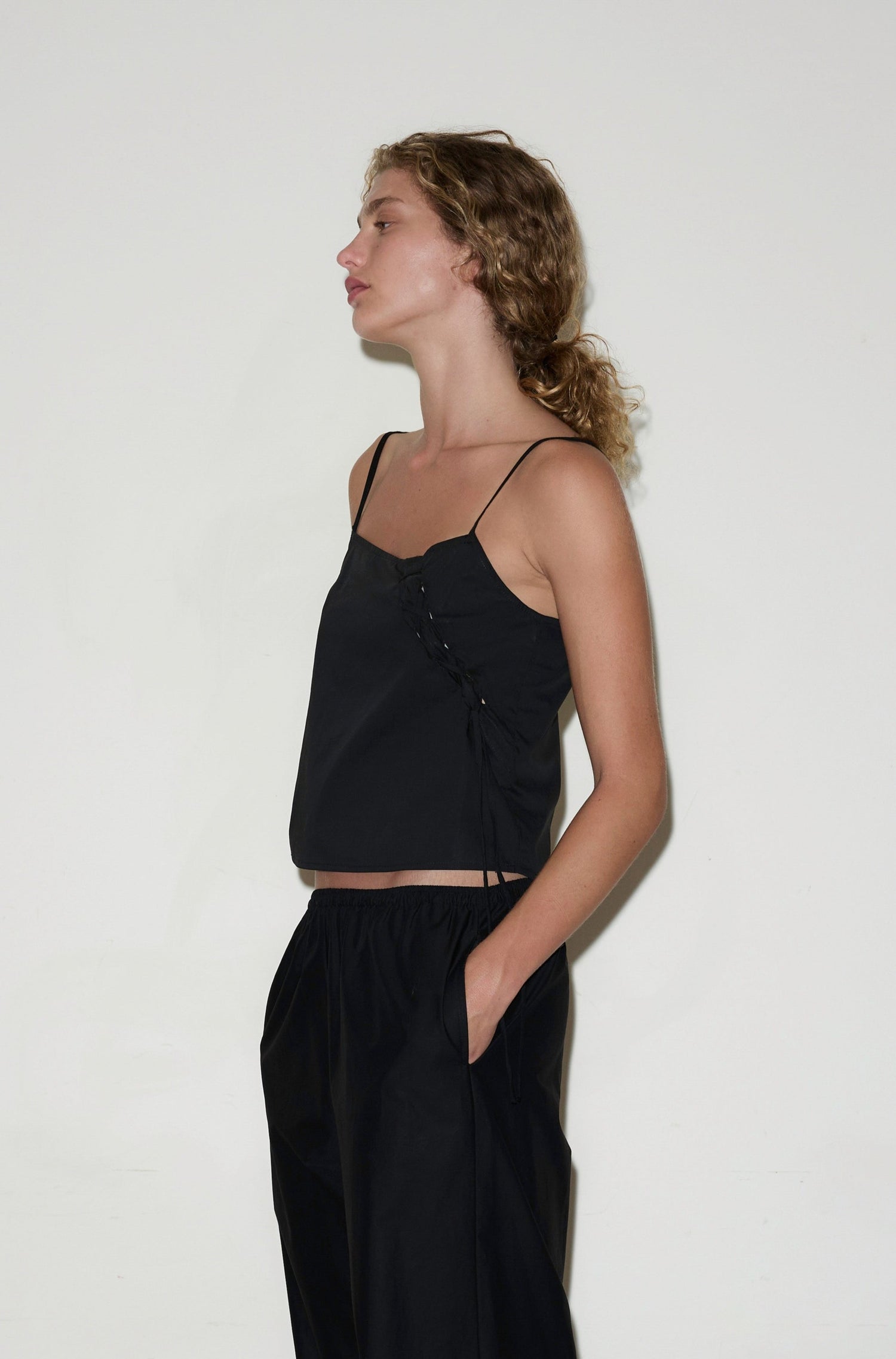 A woman with curly blonde hair in a ponytail wears the Eyelet Top - Black with thin straps and black pants, standing sideways with her hands in her pockets against a plain white background.