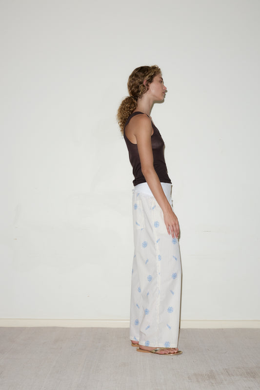 A woman stands in profile against a plain white wall, wearing a black sleeveless top and the Linen Fray belt - White by Deiji Studios as wide-leg pants with a light blue pattern. She is barefoot on a light-colored floor.