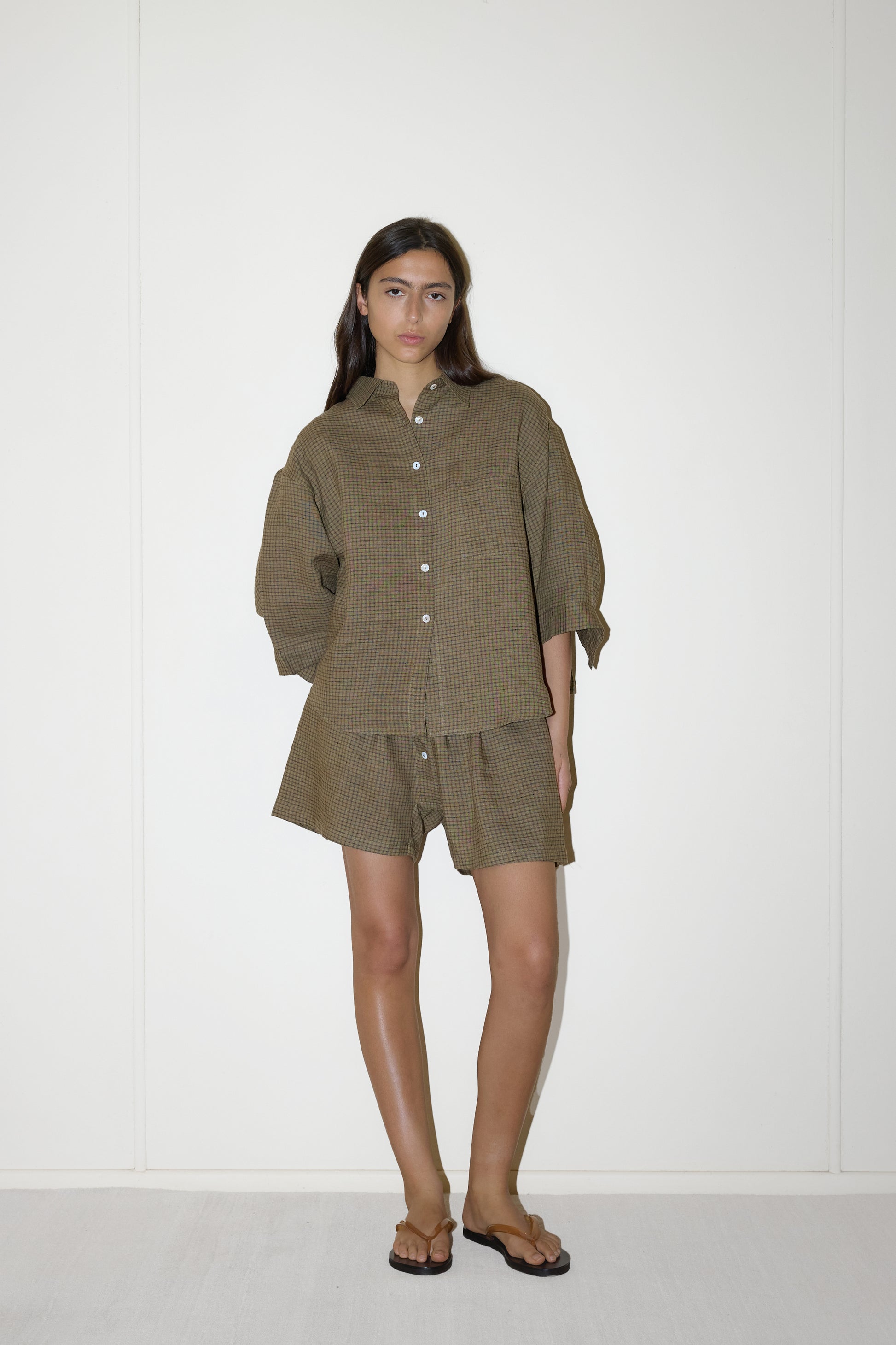 dark haired female model wears a brown and black gingham print shirt and short set in an oversized box fit linen shirt with wide arms and a front pocket designed to sit mid rise, the loose fitting boxer style shorts have a faux button down fly and an elastic waist for extra comfort