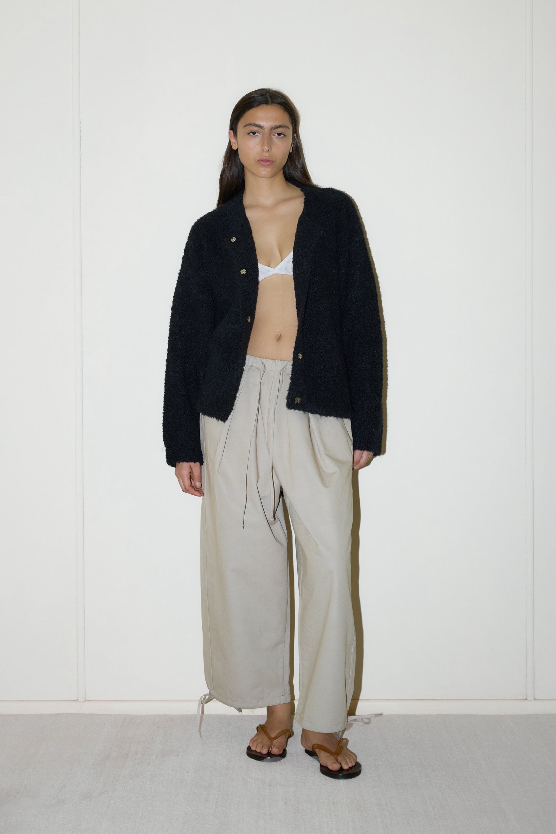 darked haired female model wears a black relaxed and textured knit features a round shaped neckline with press stud front closure, removable self tie belt with long sleeves and high back neckline, paired with cream structured cotton twill trouser featuring a drawcord hem and waist and deep side pockets designed to sit mid-low waist for a relaxed look.