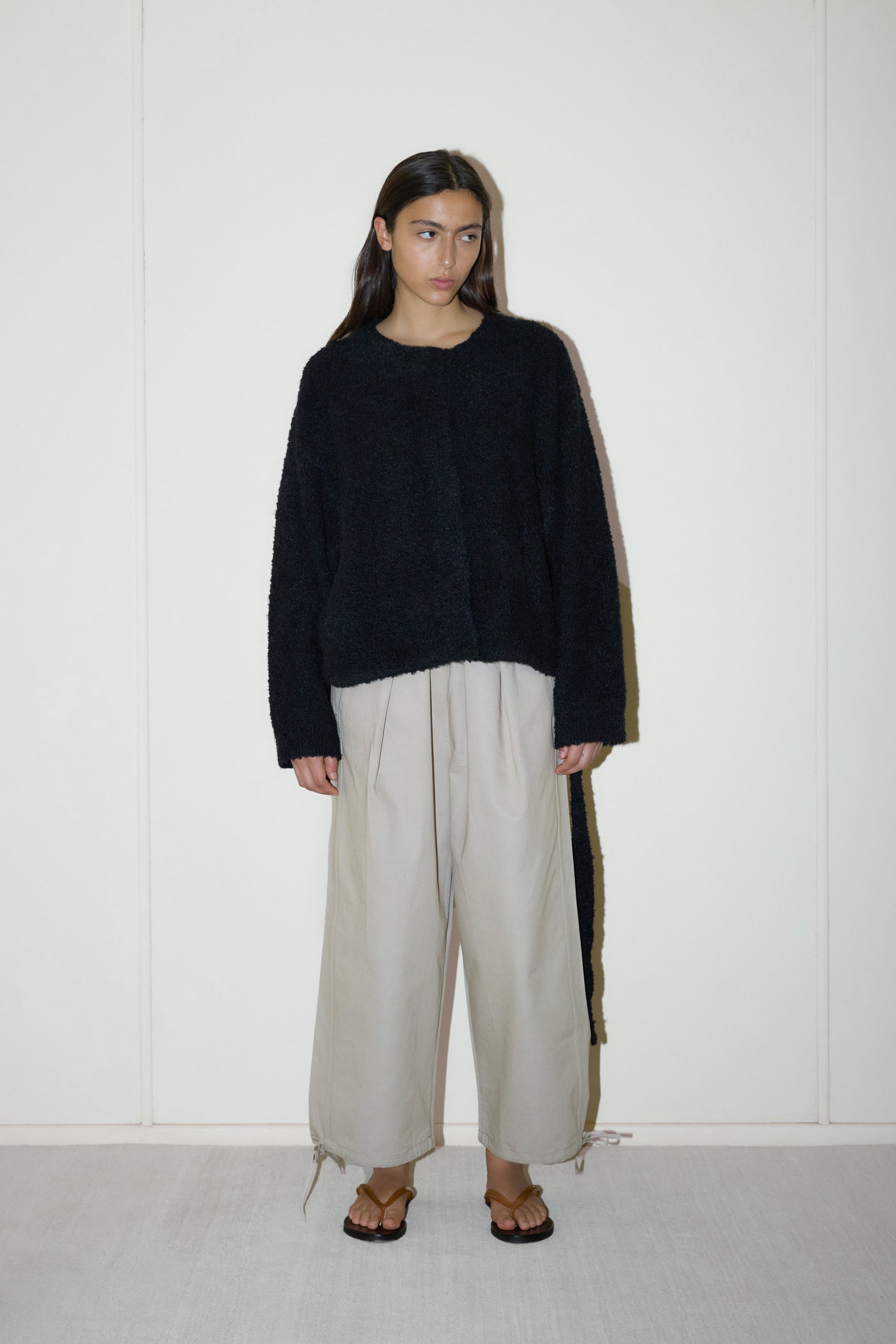 darked haired female model wears a black relaxed and textured knit features a round shaped neckline with press stud front closure, removable self tie belt with long sleeves and high back neckline, paired with cream structured cotton twill trouser featuring a drawcord hem and waist and deep side pockets designed to sit mid-low waist for a relaxed look.