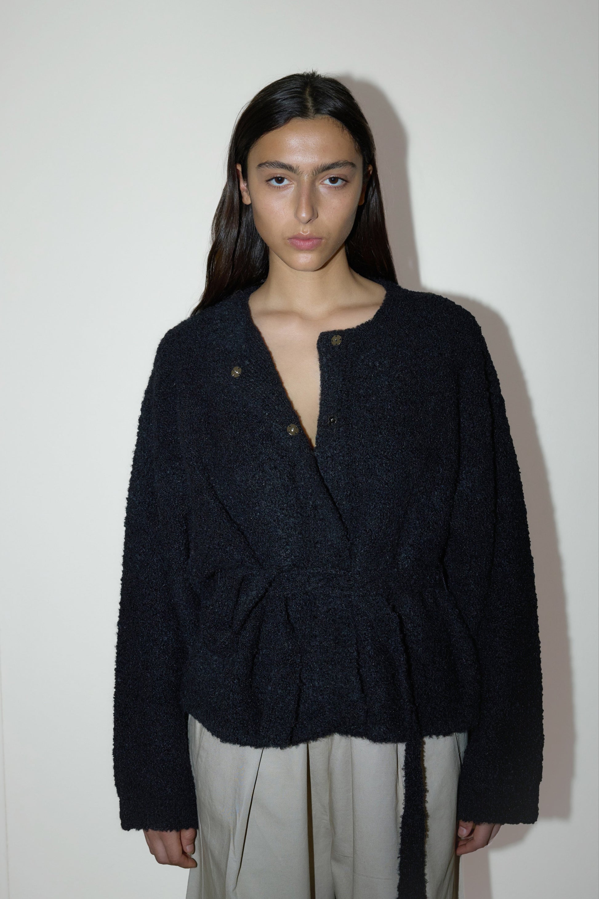 darked haired female model wears a black relaxed and textured knit features a round shaped neckline with press stud front closure, removable self tie belt with long sleeves and high back neckline, paired with cream structured cotton twill trouser featuring a drawcord hem and waist and deep side pockets designed to sit mid-low waist for a relaxed look.