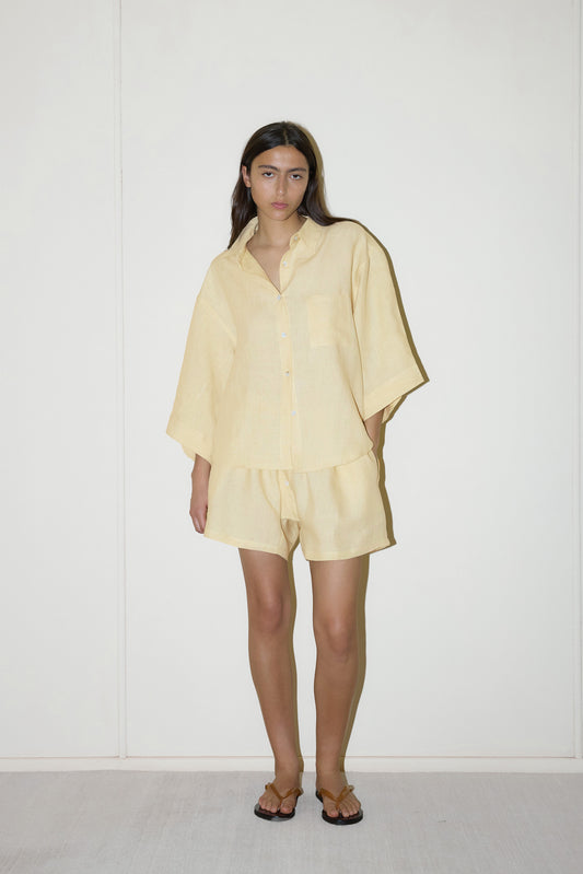 dark haired female model wears a butter yellow shirt and short set in an oversized box fit linen shirt with wide arms and a front pocket designed to sit mid rise, the loose fitting boxer style shorts have a faux button down fly and an elastic waist for extra comfort