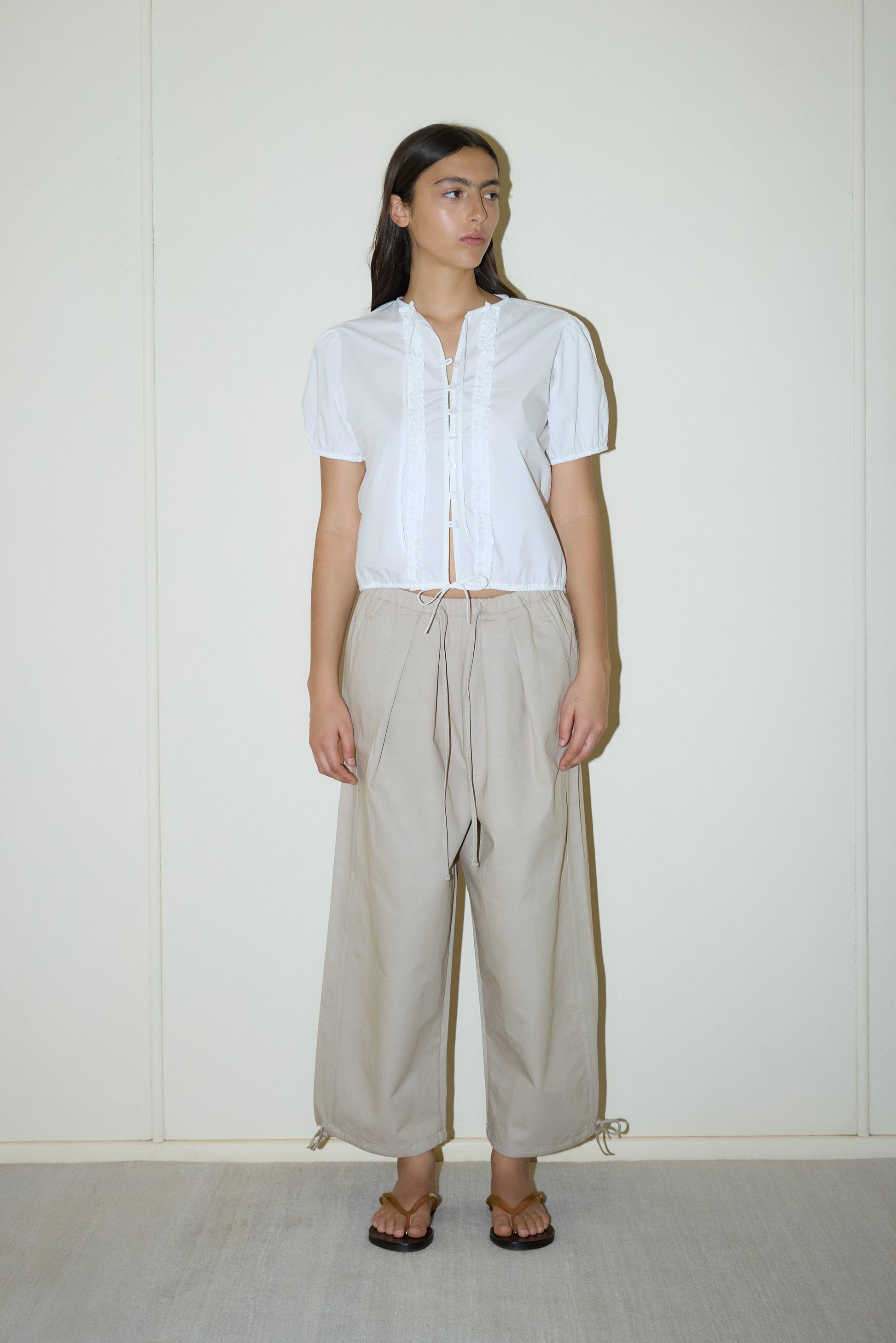 dark haired female model wears a white elevated everyday top features a flattering v neckline with button down front, fixed ruffle details, soft bloussant sleeve and gathered elastic cuffs, paired with a beige structured cotton twill trouser featuring a drawcord hem and waist and deep side pockets designed to sit mid-low waist for a relaxed look.