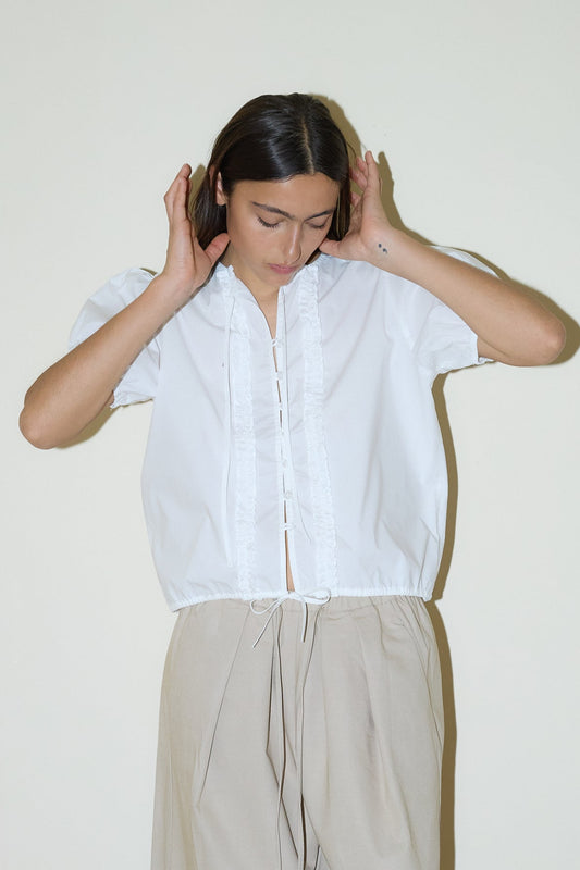 dark haired female model wears a white elevated everyday top features a flattering v neckline with button down front, fixed ruffle details, soft bloussant sleeve and gathered elastic cuffs, paired with a beige structured cotton twill trouser featuring a drawcord hem and waist and deep side pockets designed to sit mid-low waist for a relaxed look.