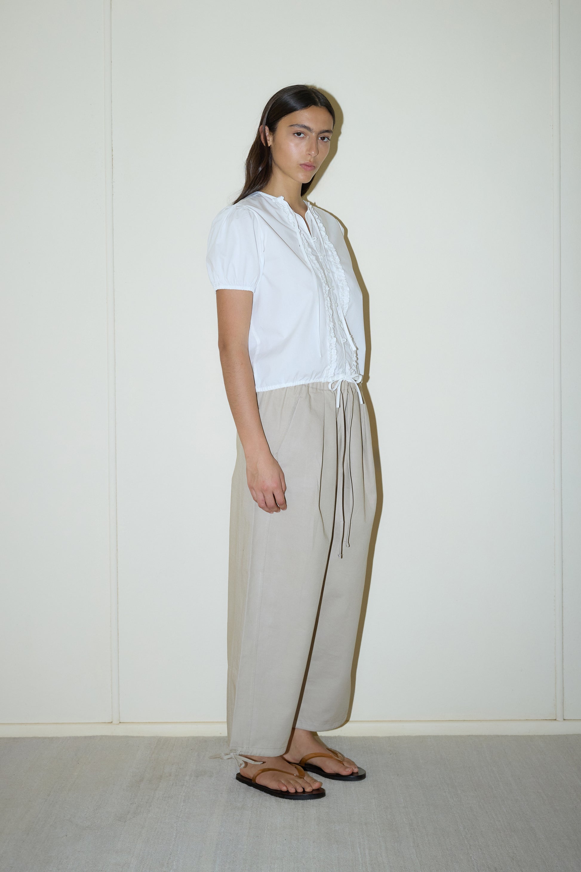 dark haired female model wears a white elevated everyday top features a flattering v neckline with button down front, fixed ruffle details, soft bloussant sleeve and gathered elastic cuffs, paired with a beige structured cotton twill trouser featuring a drawcord hem and waist and deep side pockets designed to sit mid-low waist for a relaxed look.