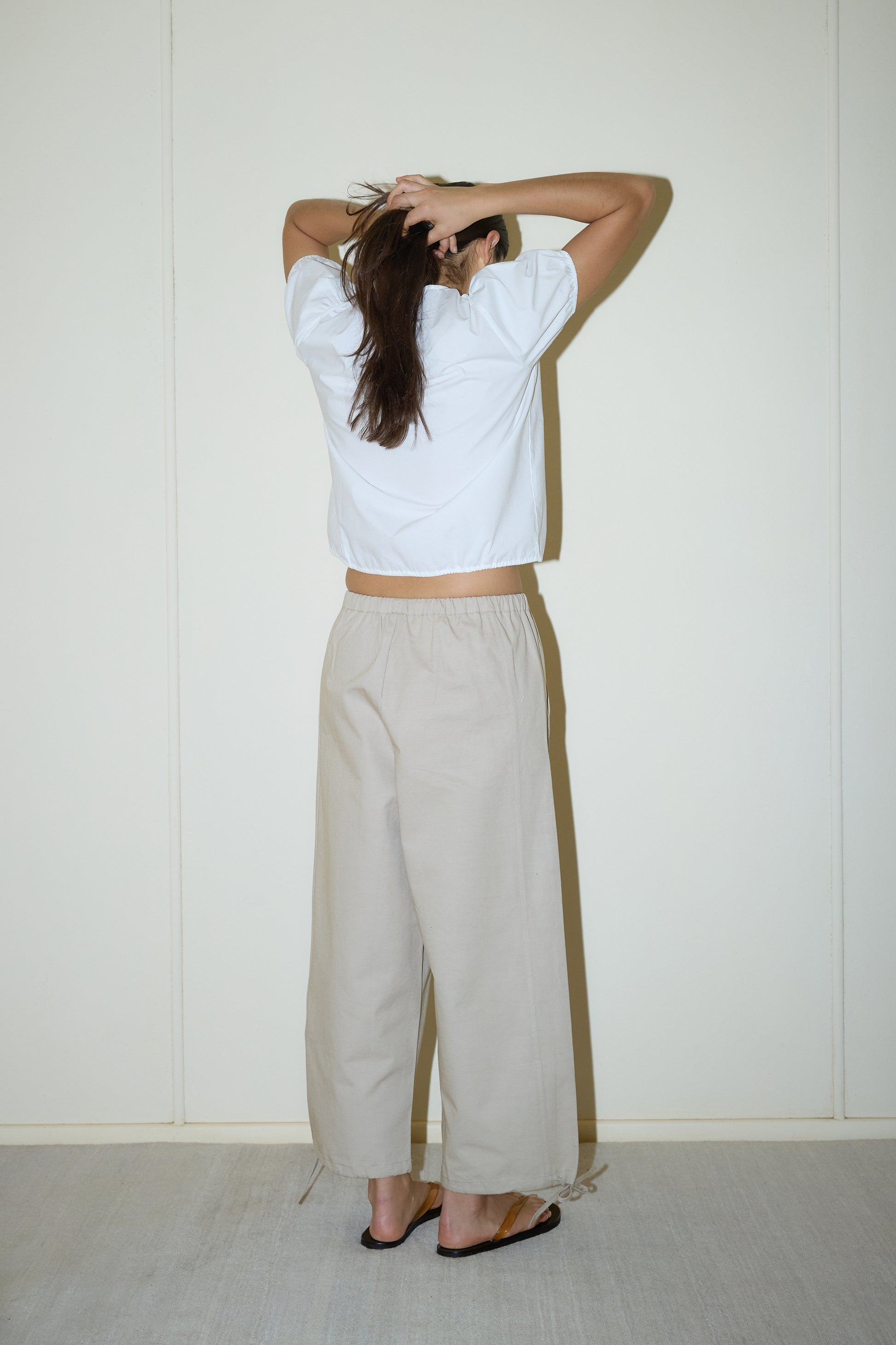 dark haired female model wears a white elevated everyday top features a flattering v neckline with button down front, fixed ruffle details, soft bloussant sleeve and gathered elastic cuffs, paired with a beige structured cotton twill trouser featuring a drawcord hem and waist and deep side pockets designed to sit mid-low waist for a relaxed look.