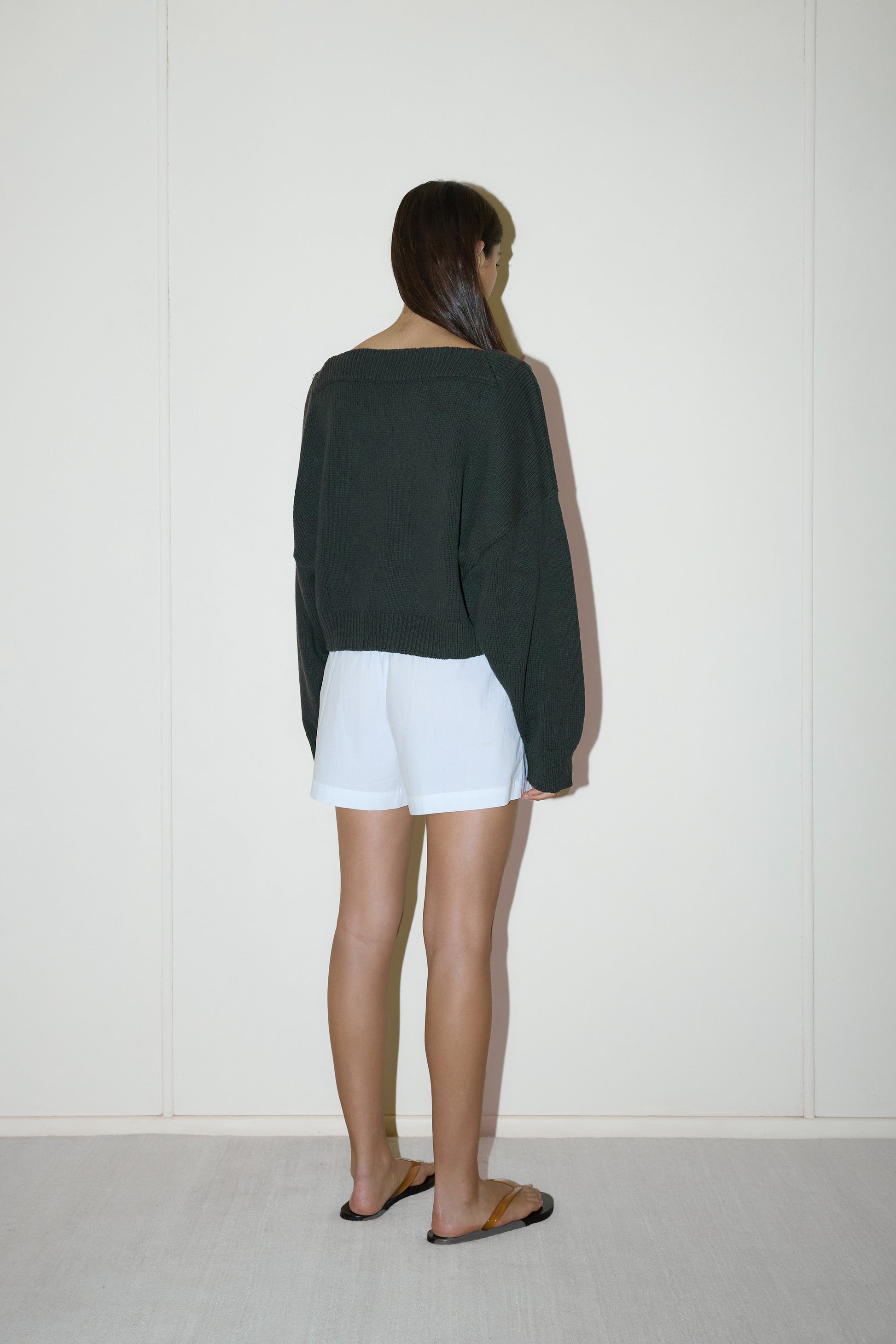 dark haired female model wears a forest green cropped, square-neck cardigan featuring snap button closure at front designed with a raised neckline for added structure and ribbed detailing at the cuffs and hem, paired with white easy pull-on boxer short designed to fit mid-rise, features a gathered elastic waistline with double button detailing.