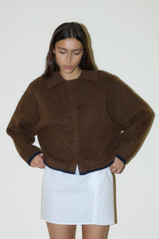 dark haired female model wears a brown elevated in a luxurious softly weighted and subtly lustrous sustainable knit blend featuring invisible press stud closure front, a relaxed collar and dropped shoulder, a gently curved arm for soft shaping and oversized fit for layering, paired with a white simple mini skirt designed to sit at low waist features adjustable tie and ruffle edge on side with button detail