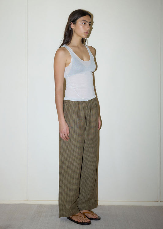 A person stands against a plain, light wall wearing a white sleeveless tank top, Deiji Studios' Linen Ease Trouser in Pine Check, and black flip-flops. They have long dark hair and a neutral expression, looking slightly to the side.