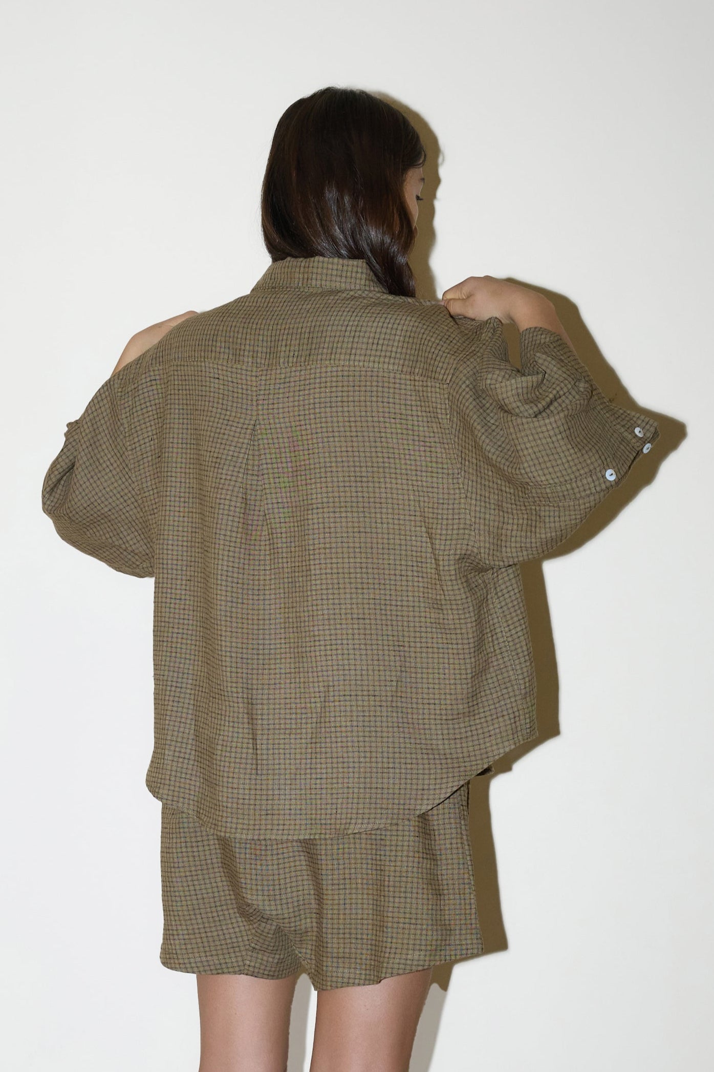 dark haired female model wears a brown and black gingham print shirt and short set in an oversized box fit linen shirt with wide arms and a front pocket designed to sit mid rise, the loose fitting boxer style shorts have a faux button down fly and an elastic waist for extra comfort