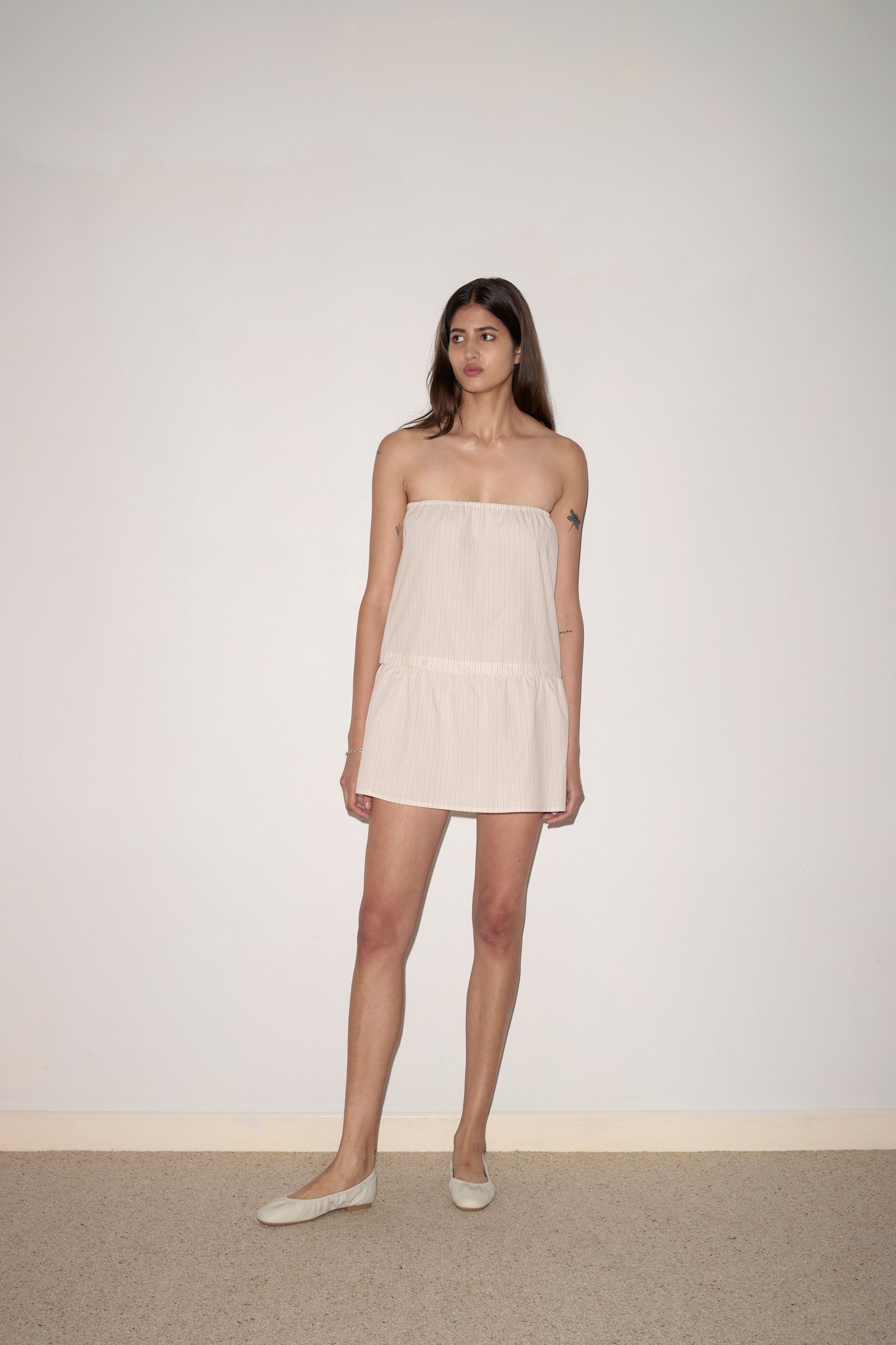 A woman with long brown hair stands against a white wall, wearing the Button Dress - Dusty Stripe. The strapless mini dress in light beige is paired with matching flats as she looks to the side with a neutral expression.