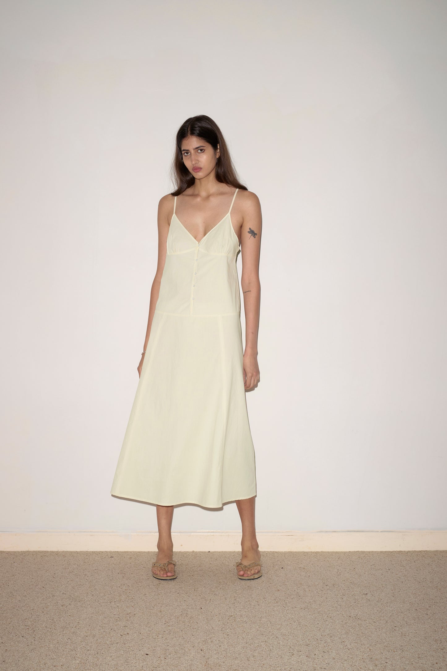 A woman with long brown hair stands against an off-white wall, wearing the Double Seam Dress - Butter Yellow by Deiji Studios with thin straps and beige sandals. She has a neutral expression and a star tattoo on her upper arm.