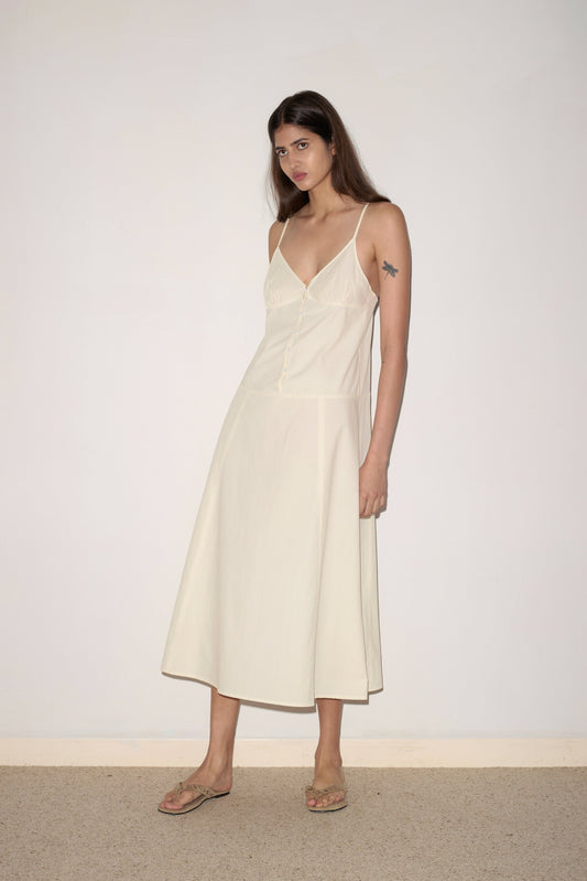 A woman with long brown hair stands against a plain white wall wearing the 0 Double Seam Dress - Butter White, a sleeveless organic cotton midi with spaghetti straps and double seams, paired with beige sandals. She has a star tattoo on her upper left arm.