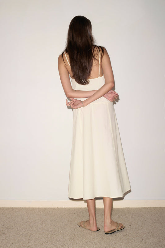 A woman with long brown hair wears the 0 Double Seam Dress in Butter White, standing with her arms crossed behind her back, facing a plain white wall.