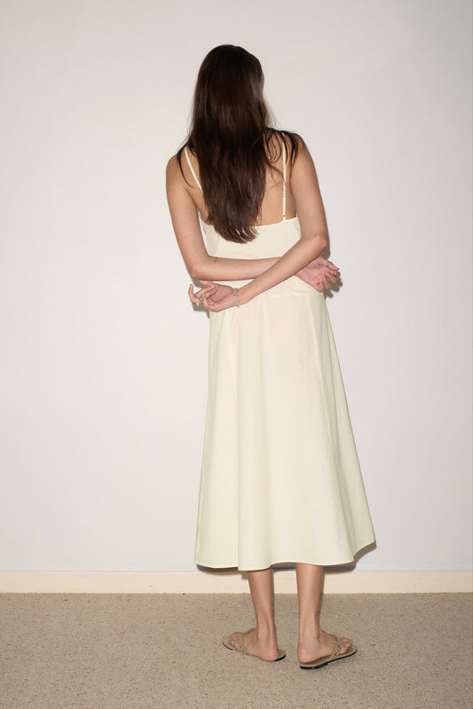 A woman with long brown hair stands facing a plain white wall, wearing the Double Seam Dress in butter yellow by Deiji Studios, her arms crossed behind her back and sandals on her feet.