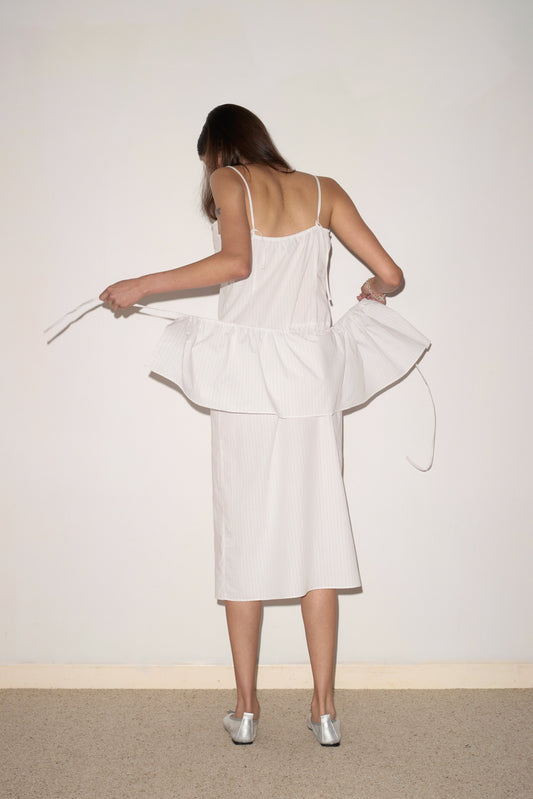 A woman with long brown hair, wearing the Peplum Dress - Pacific Stripe by 0 and silver shoes, stands facing a blank wall and adjusts the peplum detail. Her face is not visible.