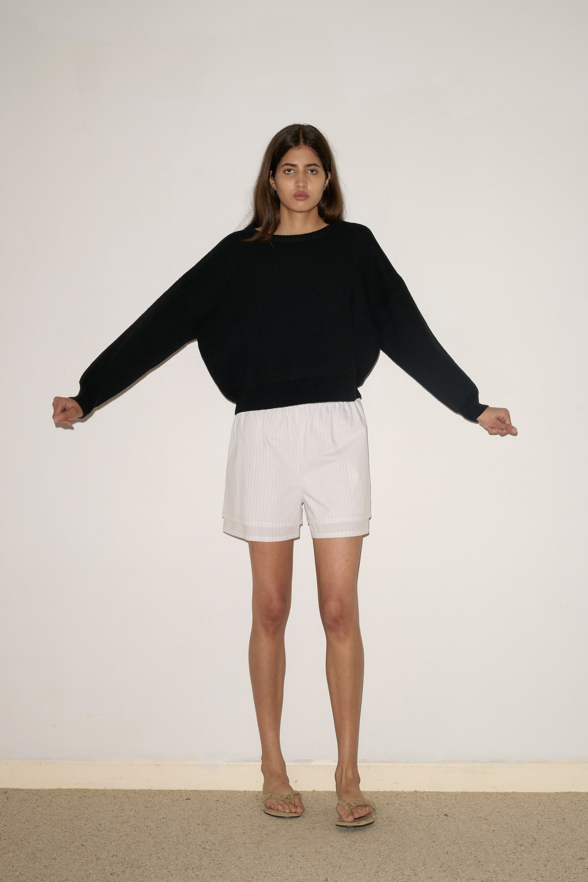 A woman stands against a plain white wall with her arms slightly outstretched, wearing the 0 Pullover - Black, white shorts, and sandals.
