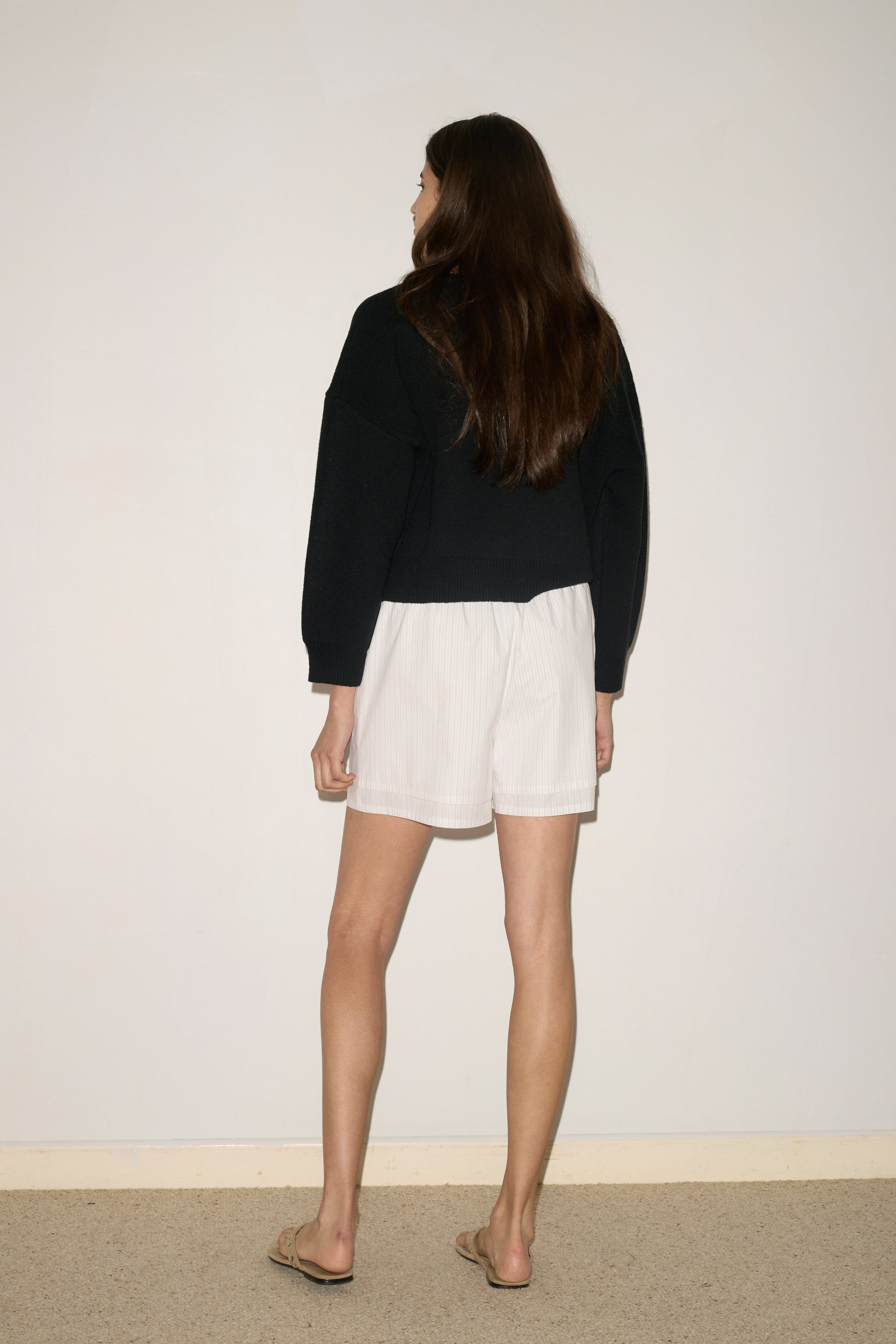 A woman with long brown hair stands facing a plain light wall, wearing a 0 Pullover - Black, white shorts, and flip-flops. Her arms are relaxed at her sides.