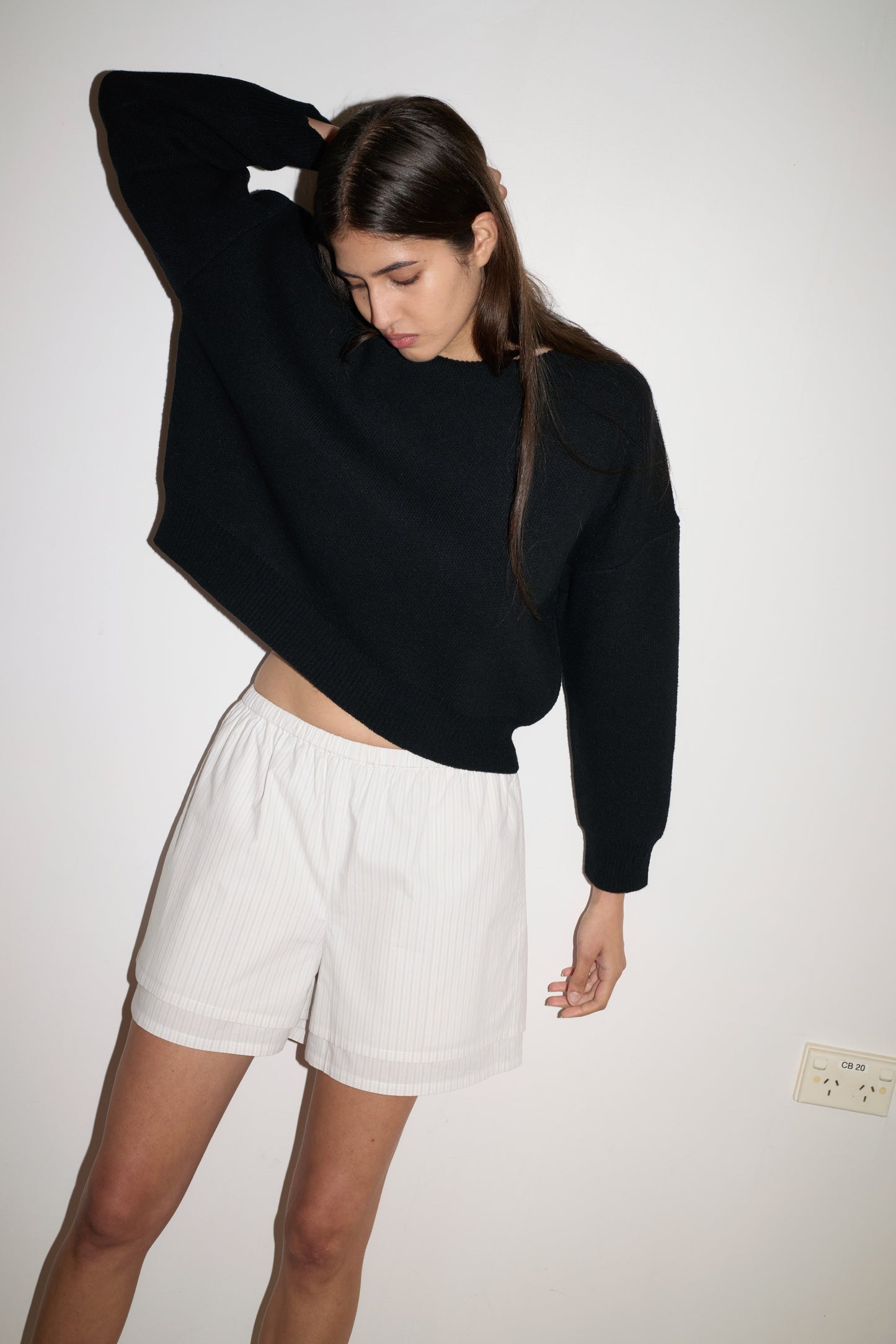 A woman stands against a plain white wall wearing the Pullover - Black from 0 and white shorts. She has long brown hair, looks down, and bends one arm behind her head.