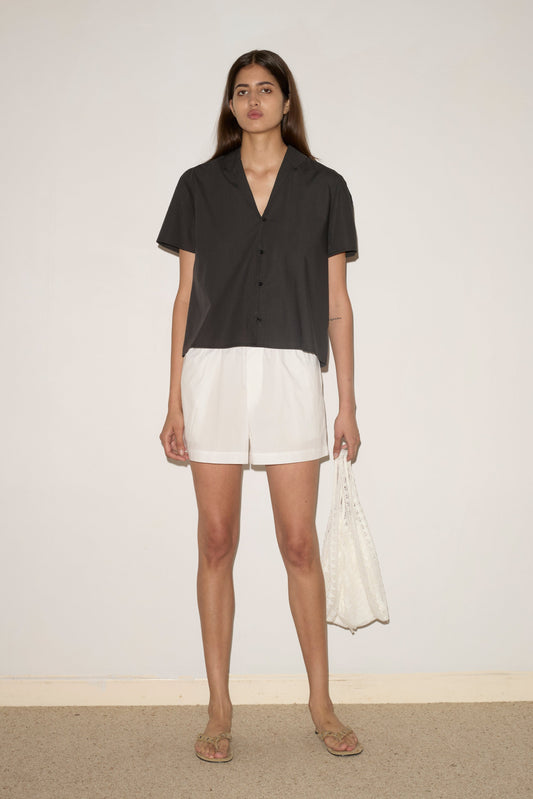 A woman with straight brown hair and a neutral expression stands against a plain wall wearing the Sails Top - Black Stripe, white shorts, flip-flops, and holding a textured white tote bag.