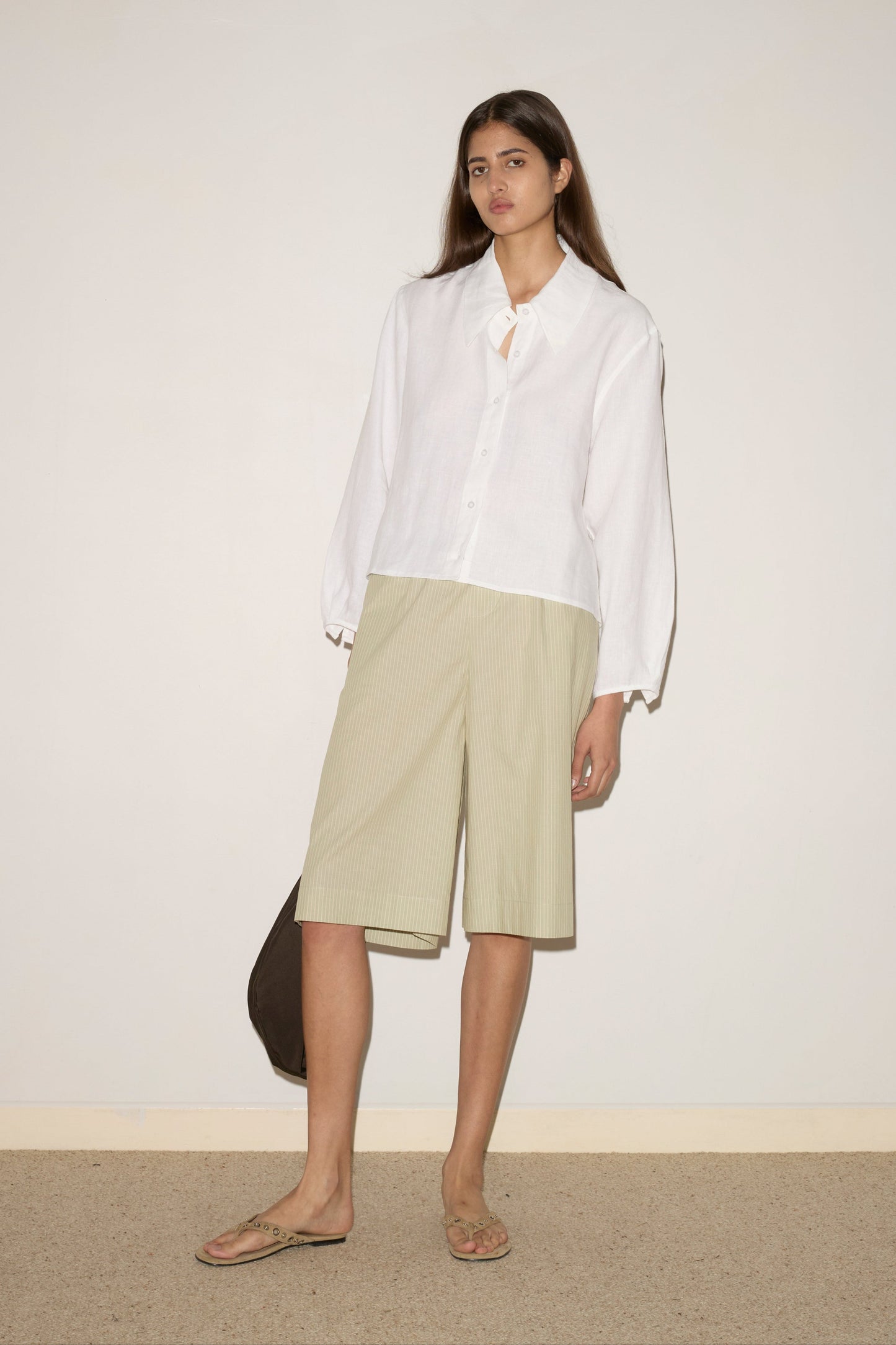 A woman with long straight hair stands against a plain wall, wearing the white Collar Point Shirt, beige knee-length shorts, flip-flops, and holding a dark bag. She has a neutral expression.