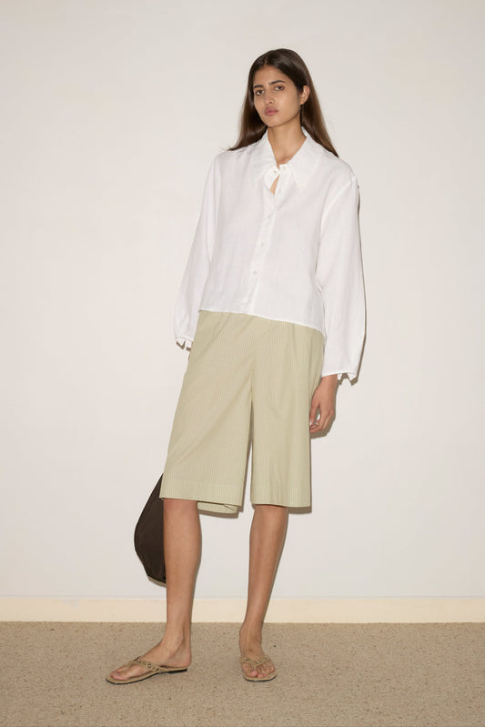 A woman with long straight hair stands against a plain wall, wearing the white Collar Point Shirt, beige knee-length shorts, flip-flops, and holding a dark bag. She has a neutral expression.