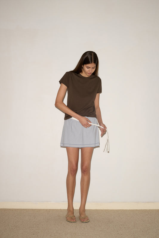 A woman with long brown hair wears the Layered Skirt - Sky Stripe, a brown organic cotton T-shirt, and sandals as she looks down to tie a white drawstring in front of a light-colored wall.