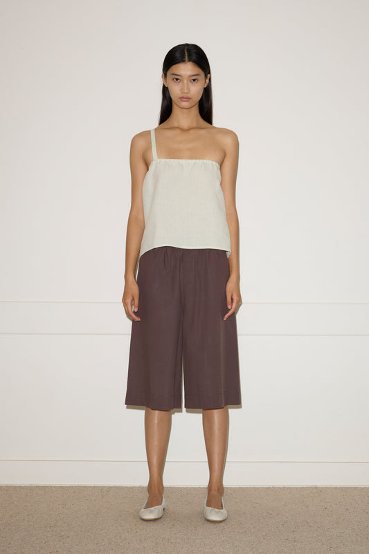 A woman stands against a light wall, wearing the Button Strap Top in Pastel Pistachio, loose brown culotte shorts, and beige flats. She faces forward with a neutral expression and straight dark hair.