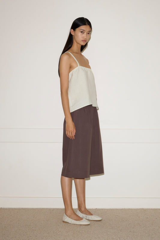 A woman with long dark hair stands sideways against a plain white wall, wearing the Button Strap Top in Pastel Pistachio, loose brown culottes, and light-colored flat shoes. She looks toward the camera with a neutral expression.