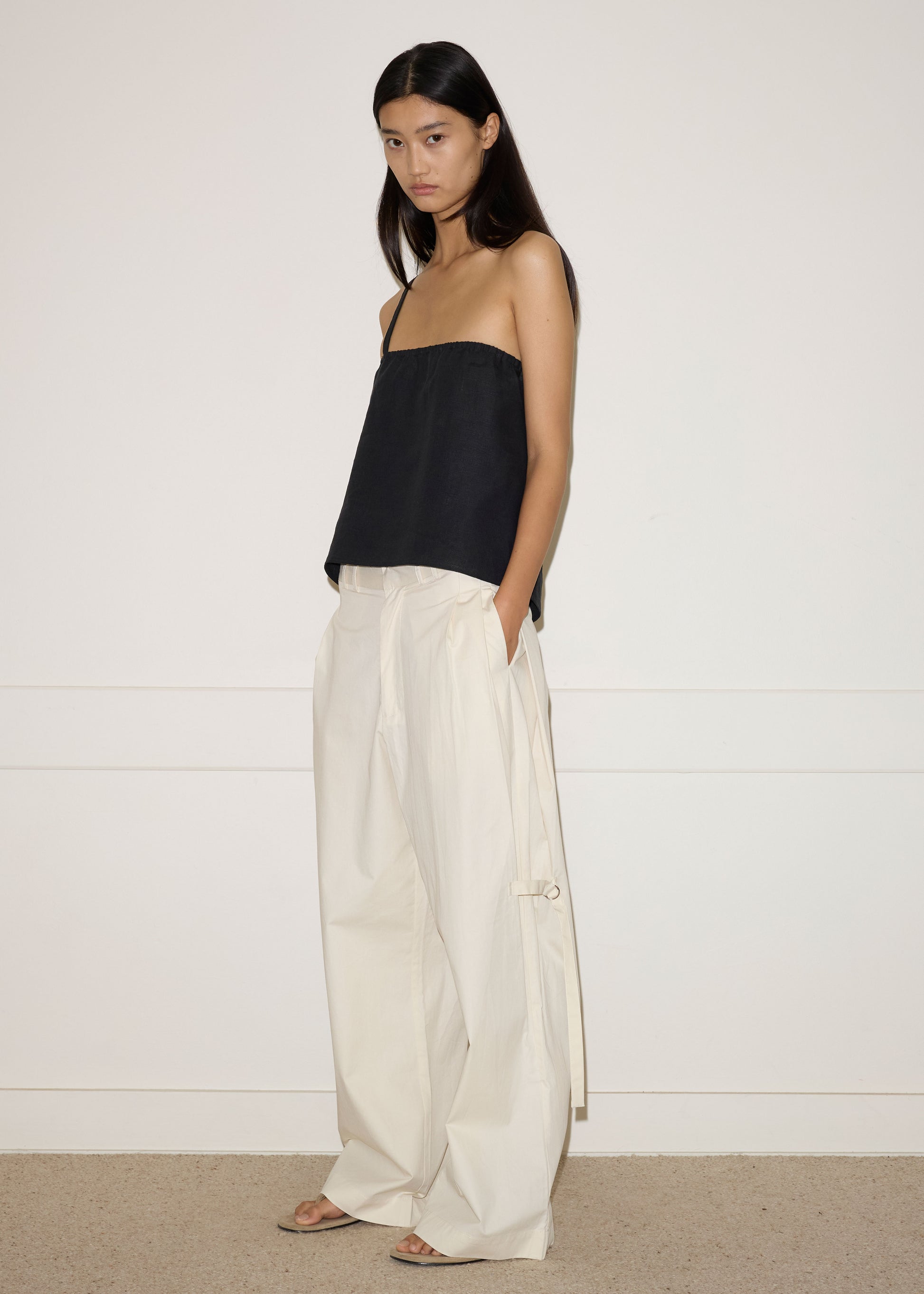 A woman stands indoors against a plain white wall, wearing a black strapless top and Deiji Studios’ Tab Pant in off white—wide-leg, high-waisted organic cotton pants with side ties. She has long straight hair and a neutral expression.