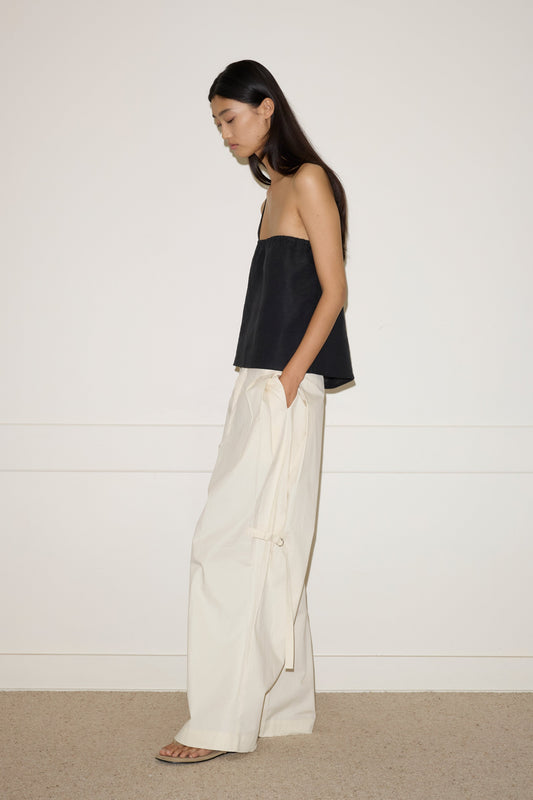 A woman stands sideways against a plain wall, wearing Deiji Studios Button Strap Top in black, loose-fitting white tie-detail trousers, and sandals. Her long dark hair is down and her hands are in her pockets.