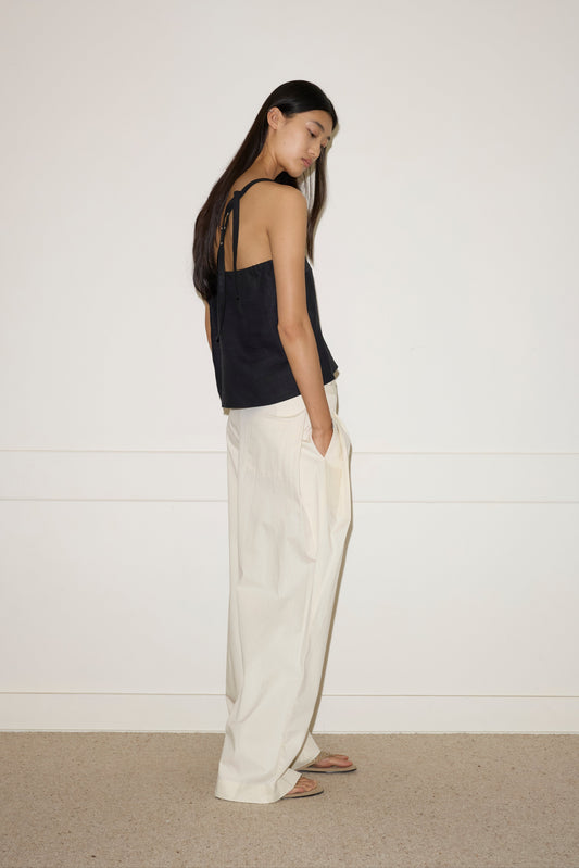 A person with long dark hair stands sideways, wearing the Deiji Studios Button Strap Top - Black with loose white linen wide-leg pants and sandals, one hand in their pocket against a plain light background.