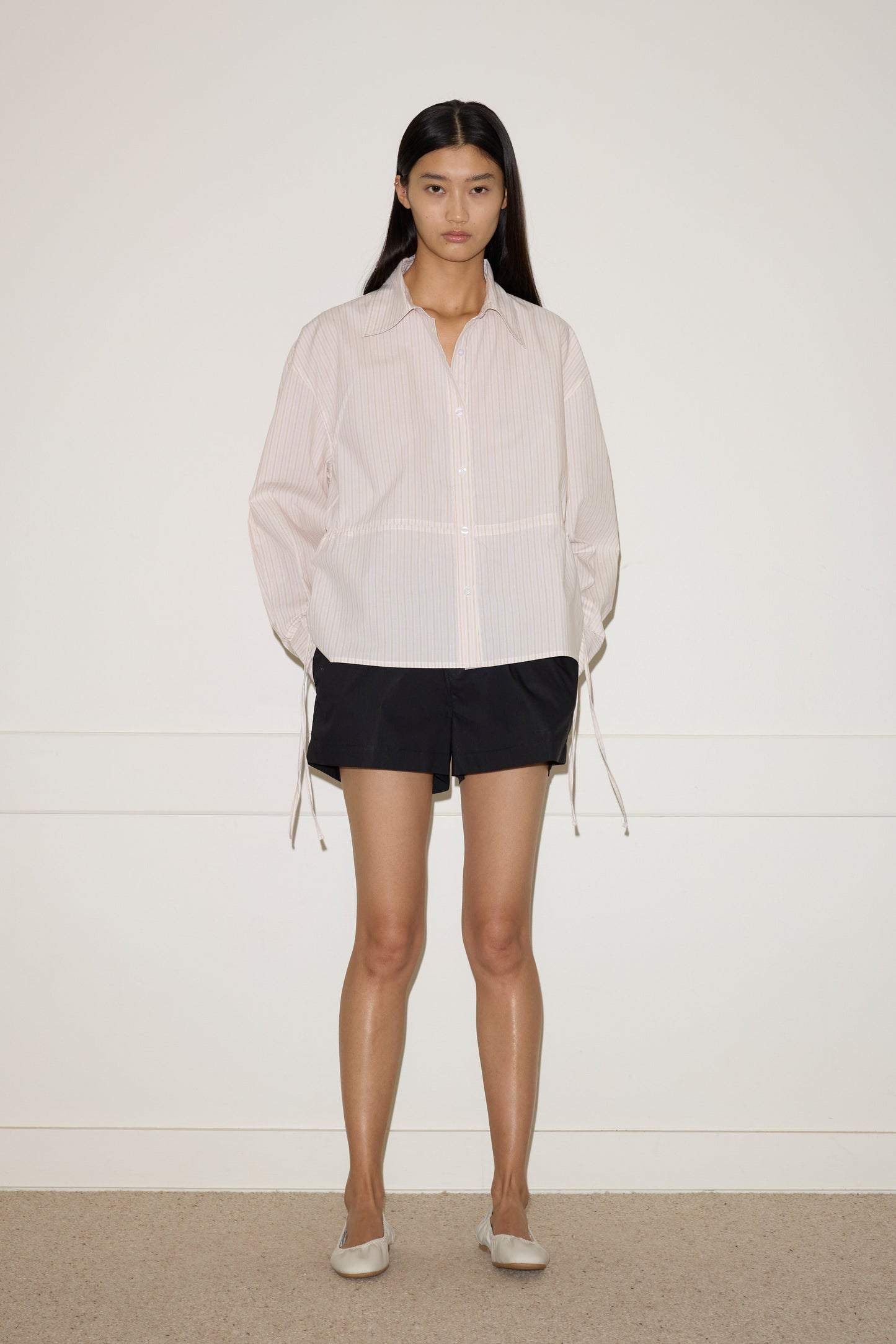 A woman with long dark hair stands against a plain white wall, wearing the Deiji Studios Room Shirt - Dusty Stripe with loose sleeves, black shorts, and light-colored flat shoes.