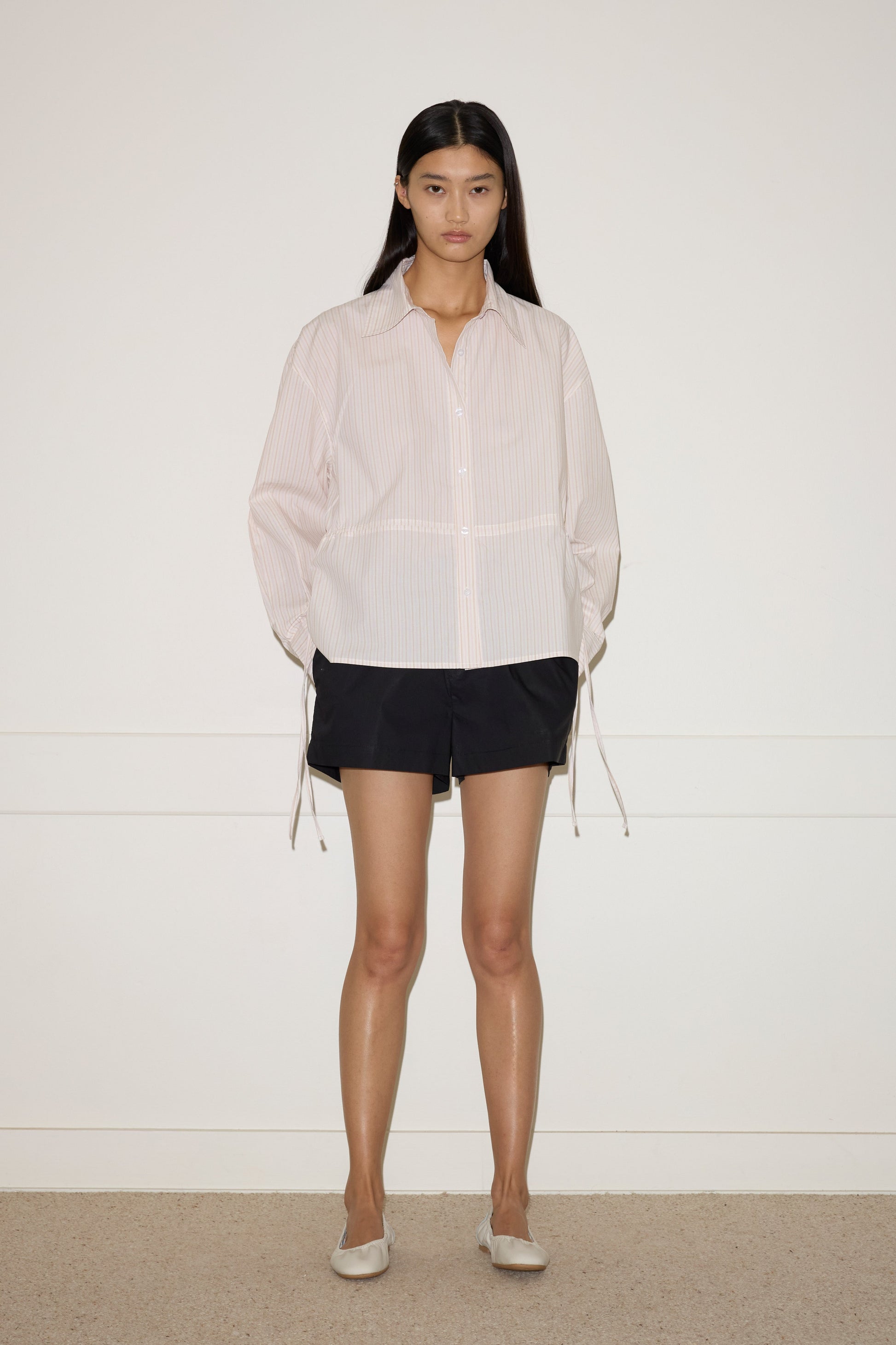 A woman with long dark hair stands against a plain white wall, wearing the Deiji Studios Room Shirt - Dusty Stripe with loose sleeves, black shorts, and light-colored flat shoes.