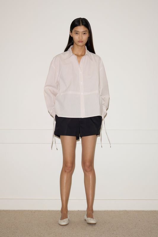 A woman with long dark hair stands against a plain white wall, wearing the Deiji Studios Room Shirt - Dusty Stripe with loose sleeves, black shorts, and light-colored flat shoes.