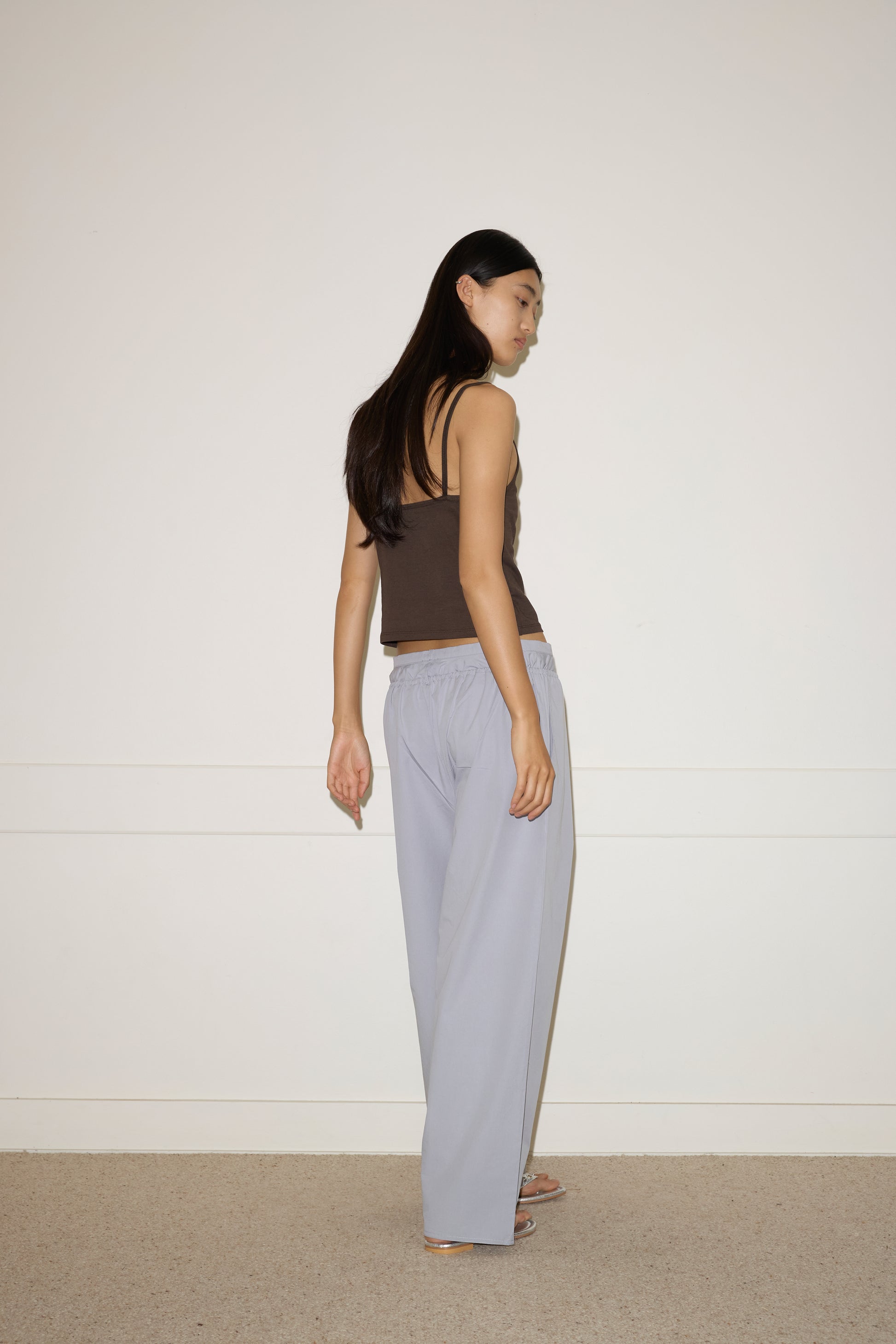 A woman with long dark hair, wearing the 0 Simple Tank - Caribou and light gray loose pants, stands in a minimalist room with a white wall and beige carpet, looking over her shoulder.